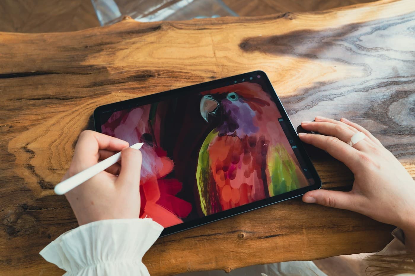 An artist painting a vivid parrot on a tablet with a stylus, resting on a wooden table.